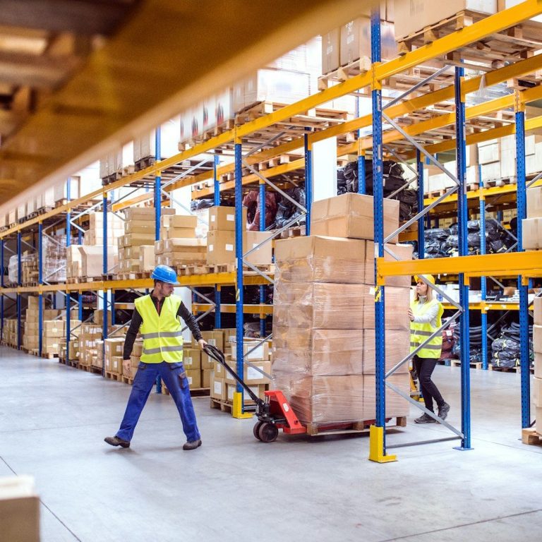 Workers in a warehouse moving boxes with a pallet truck between shelves.
